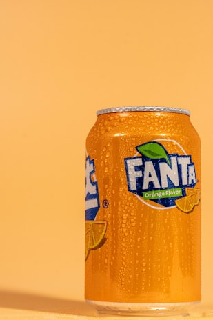 A can of orange soda is positioned against a solid orange background. The can is covered in droplets of condensation, emphasizing its cold and refreshing quality. The vibrant branding features a blue and green logo with slices of oranges, indicating the drink's flavor.