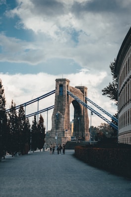 A large stone suspension bridge with blue steel cables is situated in an urban environment. The sky is overcast with scattered clouds. A group of people is gathered near the bridge, and trees line the pedestrian path leading to the bridge.