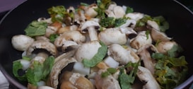A close-up view of a pan filled with fresh mushrooms and chopped green herbs. The mushrooms appear to be button mushrooms, mixed with pieces of leafy herbs, likely parsley or cilantro, creating a fresh and earthy appeal.