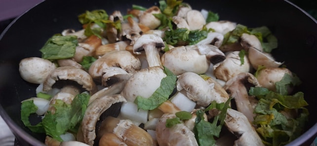 A close-up view of a pan filled with fresh mushrooms and chopped green herbs. The mushrooms appear to be button mushrooms, mixed with pieces of leafy herbs, likely parsley or cilantro, creating a fresh and earthy appeal.