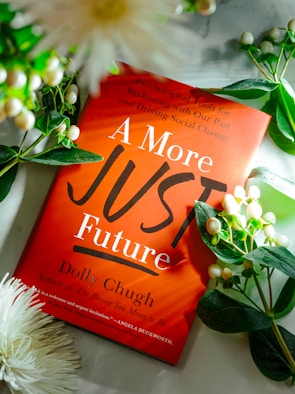 A bright red book titled 'A More Just Future' by Dolly Chugh is surrounded by greenery and white flowers. The cover includes smaller text about psychological tools for reckoning with the past and driving social change.