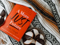 A red book titled 'A More Just Future' by Dolly Chugh is placed on a patterned textile background. Next to the book, there is a plate with several black and white cookies.