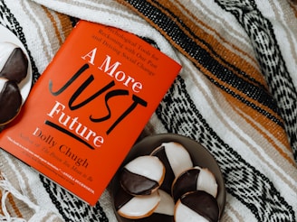 A red book titled 'A More Just Future' by Dolly Chugh is placed on a patterned textile background. Next to the book, there is a plate with several black and white cookies.