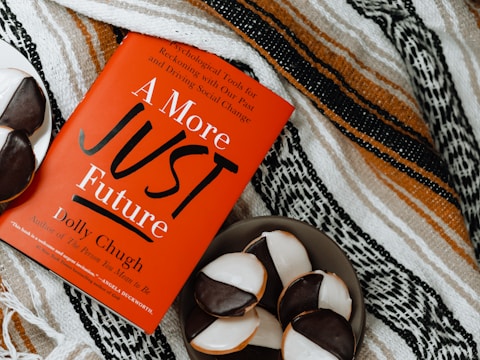 A red book titled 'A More Just Future' by Dolly Chugh is placed on a patterned textile background. Next to the book, there is a plate with several black and white cookies.