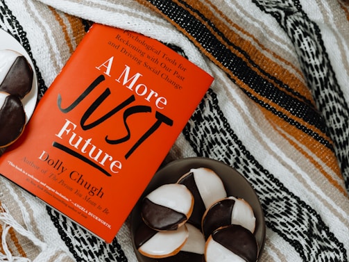 A red book titled 'A More Just Future' by Dolly Chugh is placed on a patterned textile background. Next to the book, there is a plate with several black and white cookies.