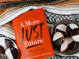 A red book titled "A More Just Future" by Dolly Chugh is placed next to a plate of black and white cookies. The book has text describing it as a tool for psychological and social change. The background includes a striped woven fabric in earthy tones.