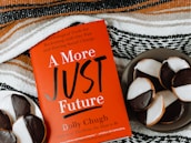 A red book titled "A More Just Future" by Dolly Chugh is placed next to a plate of black and white cookies. The book has text describing it as a tool for psychological and social change. The background includes a striped woven fabric in earthy tones.