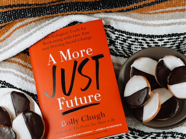 A red book titled "A More Just Future" by Dolly Chugh is placed next to a plate of black and white cookies. The book has text describing it as a tool for psychological and social change. The background includes a striped woven fabric in earthy tones.