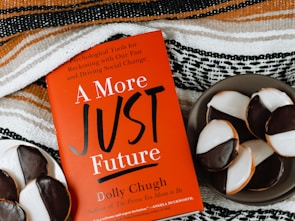 A red book titled "A More Just Future" by Dolly Chugh is placed next to a plate of black and white cookies. The book has text describing it as a tool for psychological and social change. The background includes a striped woven fabric in earthy tones.