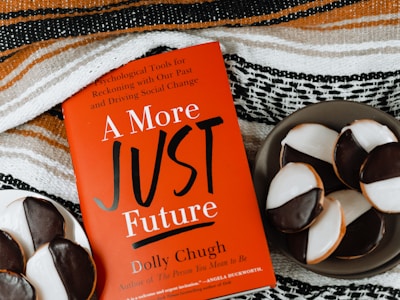 A red book titled "A More Just Future" by Dolly Chugh is placed next to a plate of black and white cookies. The book has text describing it as a tool for psychological and social change. The background includes a striped woven fabric in earthy tones.