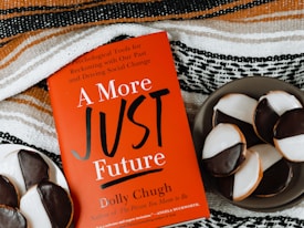 A red book titled "A More Just Future" by Dolly Chugh is placed next to a plate of black and white cookies. The book has text describing it as a tool for psychological and social change. The background includes a striped woven fabric in earthy tones.
