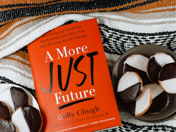 A red book titled "A More Just Future" by Dolly Chugh is placed next to a plate of black and white cookies. The book has text describing it as a tool for psychological and social change. The background includes a striped woven fabric in earthy tones.