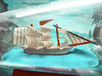 A small, detailed model ship with multiple sails is encased in a clear glass-like container. The sails are intricately designed with thin, dark lines and are set against a bright blue background that resembles the ocean. There are subtle reflections and a soft light, creating a sense of depth.