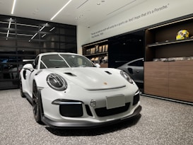 A white sports car with sleek, aerodynamic design is parked in a modern showroom with polished, speckled flooring. The car features prominent headlights, a large front grille, and dark rims. Wall displays include models of cars and racing helmets. There's a famous quote on the wall in the background near the ceiling.