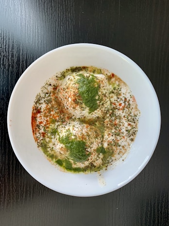Two savory dumplings sit in a white bowl, covered with creamy yogurt. They are garnished with green chutney, red chili powder, and dried herbs. The dish is presented against a dark, glossy table.