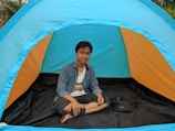 A person is sitting cross-legged inside a colorful outdoor tent. The tent has bright hues of blue and orange, and it is set up in a natural environment with greenery visible in the background. The person is casually dressed and holding a smartphone in one hand, appearing relaxed.
