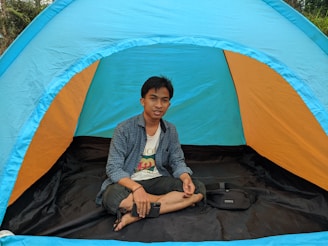 A person is sitting cross-legged inside a colorful outdoor tent. The tent has bright hues of blue and orange, and it is set up in a natural environment with greenery visible in the background. The person is casually dressed and holding a smartphone in one hand, appearing relaxed.