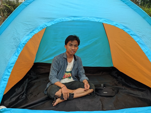 A person is sitting cross-legged inside a colorful outdoor tent. The tent has bright hues of blue and orange, and it is set up in a natural environment with greenery visible in the background. The person is casually dressed and holding a smartphone in one hand, appearing relaxed.