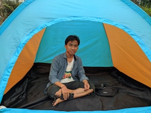 A person is sitting cross-legged inside a colorful outdoor tent. The tent has bright hues of blue and orange, and it is set up in a natural environment with greenery visible in the background. The person is casually dressed and holding a smartphone in one hand, appearing relaxed.