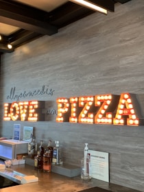 A wall display features the phrase 'all you need is LOVE and PIZZA' with illuminated marquee-style letters. Below is a countertop with various bottles of liquor, a napkin holder, and other bar accessories. The background is a wooden textured wall.