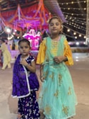 Two young girls stand together, dressed in colorful traditional attire. The younger girl on the left is wearing a purple dress with floral patterns and a bindi on her forehead. The older girl on the right is dressed in a teal dress with golden embroidery, complemented by a yellow blouse. They are standing in what seems to be a festive environment, with bright lights and decorations, including garlands and string lights in the background.