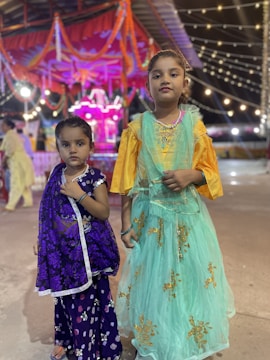 Two young girls stand together, dressed in colorful traditional attire. The younger girl on the left is wearing a purple dress with floral patterns and a bindi on her forehead. The older girl on the right is dressed in a teal dress with golden embroidery, complemented by a yellow blouse. They are standing in what seems to be a festive environment, with bright lights and decorations, including garlands and string lights in the background.