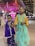 Two young girls stand together, dressed in colorful traditional attire. The younger girl on the left is wearing a purple dress with floral patterns and a bindi on her forehead. The older girl on the right is dressed in a teal dress with golden embroidery, complemented by a yellow blouse. They are standing in what seems to be a festive environment, with bright lights and decorations, including garlands and string lights in the background.