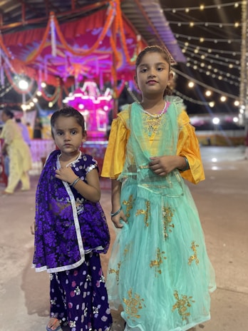 Two young girls stand together, dressed in colorful traditional attire. The younger girl on the left is wearing a purple dress with floral patterns and a bindi on her forehead. The older girl on the right is dressed in a teal dress with golden embroidery, complemented by a yellow blouse. They are standing in what seems to be a festive environment, with bright lights and decorations, including garlands and string lights in the background.