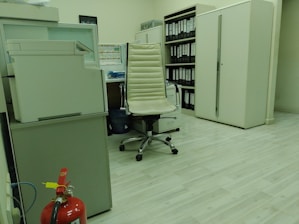 A well-organized office space with a large photocopier on the left, a modern office chair in the center, and a desk with neatly arranged blue files. Behind the chair, there is a wall of binders in a large cabinet. A fire extinguisher is on the floor near the photocopier, and the floor has a light-colored wood finish.