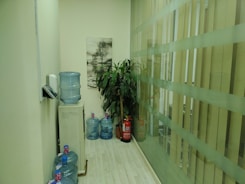 A narrow corridor with several large water dispensers placed on the light-colored wooden floor. A painting of a tree hangs on the wall above the dispensers. A tall green plant stands beside a red fire extinguisher, and there's a frosted glass partition on the right side.