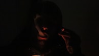 A person is holding a lit cigar or cigarette in their hand, with smoke wafting around them. The image is dark, with minimal light highlighting the glow of the cigar and the outline of the person's face.