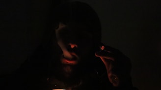 A person is holding a lit cigar or cigarette in their hand, with smoke wafting around them. The image is dark, with minimal light highlighting the glow of the cigar and the outline of the person's face.