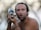 A shirtless man with a surprised or comical expression is holding a fish close to his face. His wide eyes and open mouth mimic the fish's facial features. The background is blurred, suggesting an outdoor setting.