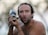 A shirtless man with a surprised or comical expression is holding a fish close to his face. His wide eyes and open mouth mimic the fish's facial features. The background is blurred, suggesting an outdoor setting.