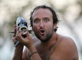 A shirtless man with a surprised or comical expression is holding a fish close to his face. His wide eyes and open mouth mimic the fish's facial features. The background is blurred, suggesting an outdoor setting.