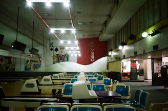 An empty bowling alley with rows of blue and white seating facing a series of lanes. Overhead lighting creates a bright atmosphere, while the walls are adorned with large playing card imagery. The red wall with the letters 'YSSC' is prominent in the background.