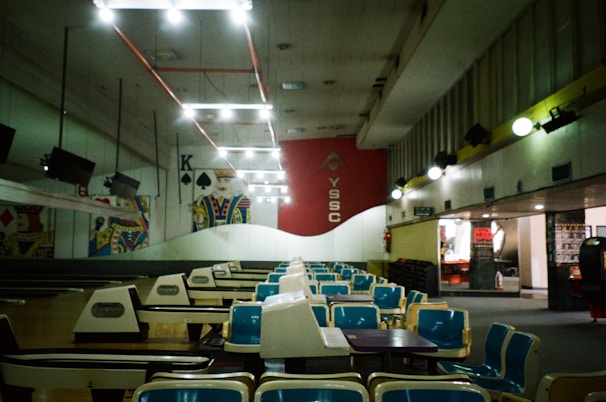 An empty bowling alley with rows of blue and white seating facing a series of lanes. Overhead lighting creates a bright atmosphere, while the walls are adorned with large playing card imagery. The red wall with the letters 'YSSC' is prominent in the background.