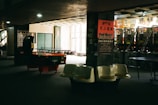 A dimly lit arcade room featuring several pinball machines lined up against a wall, with an air hockey table in the foreground. Plastic bucket seats are positioned nearby. The walls are decorated with posters containing text in a foreign language, contributing to a nostalgic or retro atmosphere.