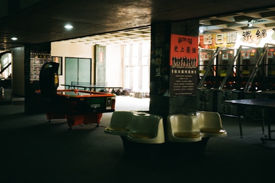 A dimly lit arcade room featuring several pinball machines lined up against a wall, with an air hockey table in the foreground. Plastic bucket seats are positioned nearby. The walls are decorated with posters containing text in a foreign language, contributing to a nostalgic or retro atmosphere.