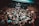 A crowded live music venue with many people gathered in front of a stage, where a band is performing. The audience, mostly dressed in dark clothing, appears engaged and excited. The environment has a grungy, underground feel with exposed brick walls and dim lighting.