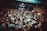 A crowded live music venue with many people gathered in front of a stage, where a band is performing. The audience, mostly dressed in dark clothing, appears engaged and excited. The environment has a grungy, underground feel with exposed brick walls and dim lighting.