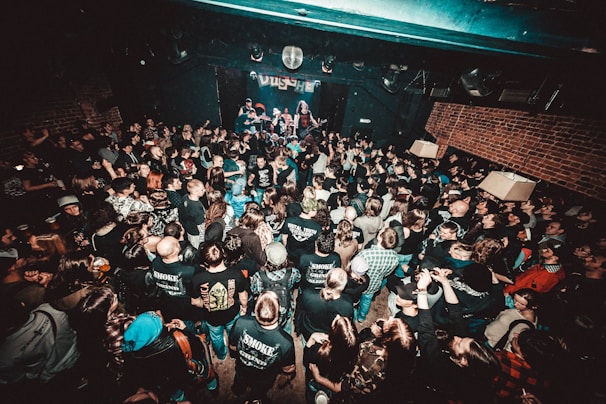 A crowded live music venue with many people gathered in front of a stage, where a band is performing. The audience, mostly dressed in dark clothing, appears engaged and excited. The environment has a grungy, underground feel with exposed brick walls and dim lighting.