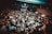 A crowded live music venue with many people gathered in front of a stage, where a band is performing. The audience, mostly dressed in dark clothing, appears engaged and excited. The environment has a grungy, underground feel with exposed brick walls and dim lighting.