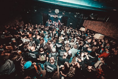 A crowded live music venue with many people gathered in front of a stage, where a band is performing. The audience, mostly dressed in dark clothing, appears engaged and excited. The environment has a grungy, underground feel with exposed brick walls and dim lighting.