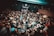 A crowded live music venue with many people gathered in front of a stage, where a band is performing. The audience, mostly dressed in dark clothing, appears engaged and excited. The environment has a grungy, underground feel with exposed brick walls and dim lighting.