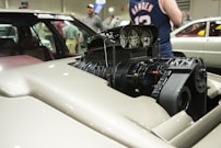 A high-performance automotive engine with a supercharger prominently displayed in the foreground. The sleek engine components are highlighted by a polished silver finish with intricate details that suggest power and precision. In the background, a crowd of people is visible, likely at a car show or exhibition, with various cars partially visible.