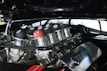 A close-up view of a car engine with custom parts, adorned with several black eight ball-style decorations and one unique red ball featuring a cartoonishly angry face. The metal components are highly polished, reflecting the surrounding environment.