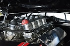 A close-up view of a car engine with custom parts, adorned with several black eight ball-style decorations and one unique red ball featuring a cartoonishly angry face. The metal components are highly polished, reflecting the surrounding environment.