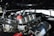 A close-up view of a car engine with custom parts, adorned with several black eight ball-style decorations and one unique red ball featuring a cartoonishly angry face. The metal components are highly polished, reflecting the surrounding environment.