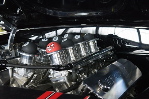A close-up view of a car engine with custom parts, adorned with several black eight ball-style decorations and one unique red ball featuring a cartoonishly angry face. The metal components are highly polished, reflecting the surrounding environment.